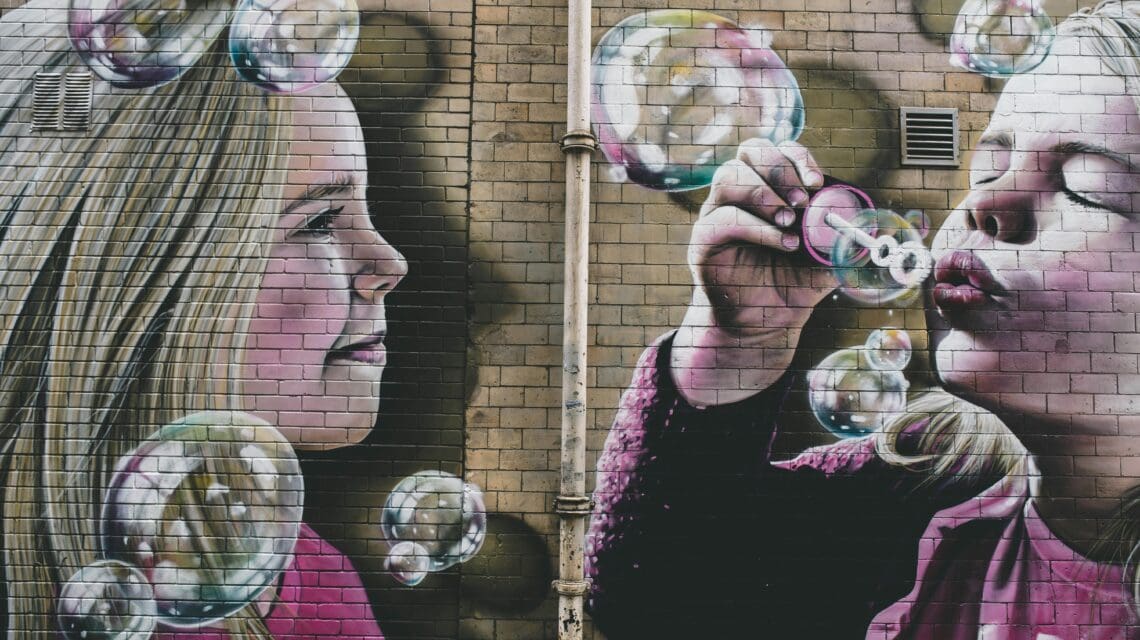 A vibrant mural on a brick wall depicts two children with expressions of joy. One blows colorful bubbles, while the other watches in awe. The scene conveys whimsy and innocence.