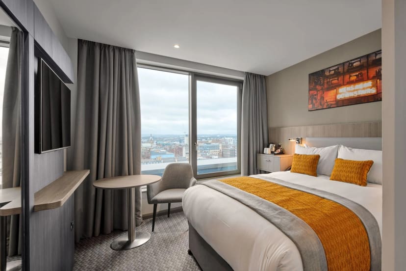 Modern hotel room with a large bed, accented by orange pillows and a gray throw. Features a city view from a large window, a wall-mounted TV, and minimalist decor.