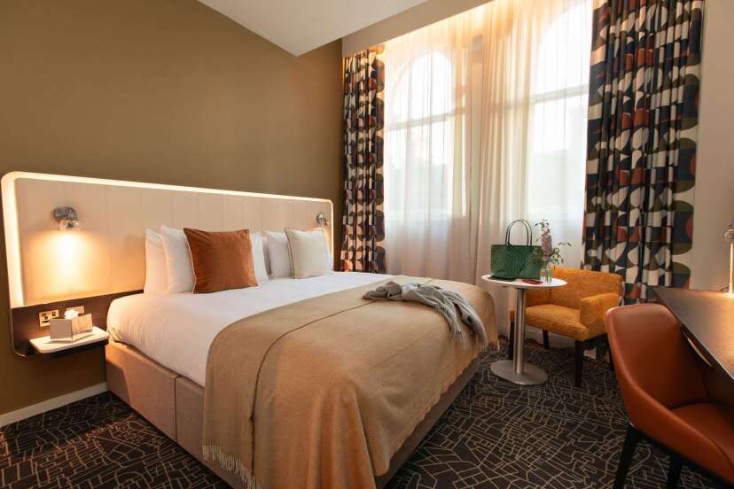 Modern hotel room featuring a cozy bed with decorative pillows, stylish armchair, and large windows with flowing curtains.