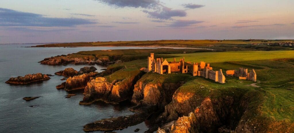 Ancient castle ruins bathed in golden sunset light atop a rocky cliff by the sea, surrounded by lush green fields and a calm, expansive ocean.