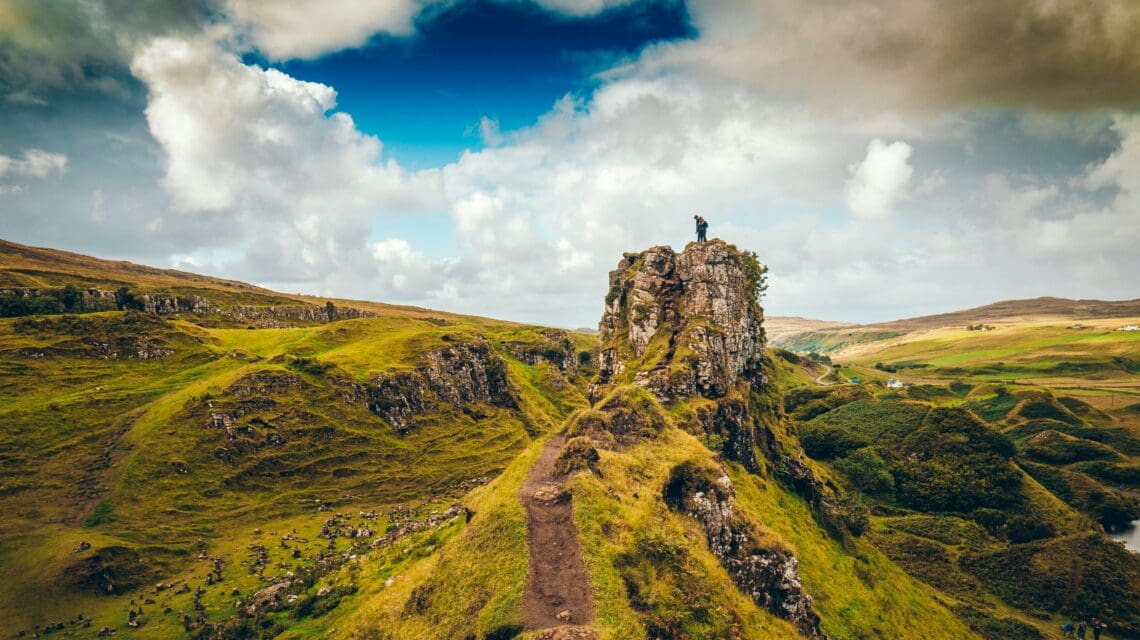 A lone person stands atop a rugged, grassy hill, overlooking a vast, rolling landscape under a partly cloudy blue sky. The scene conveys a sense of adventure.