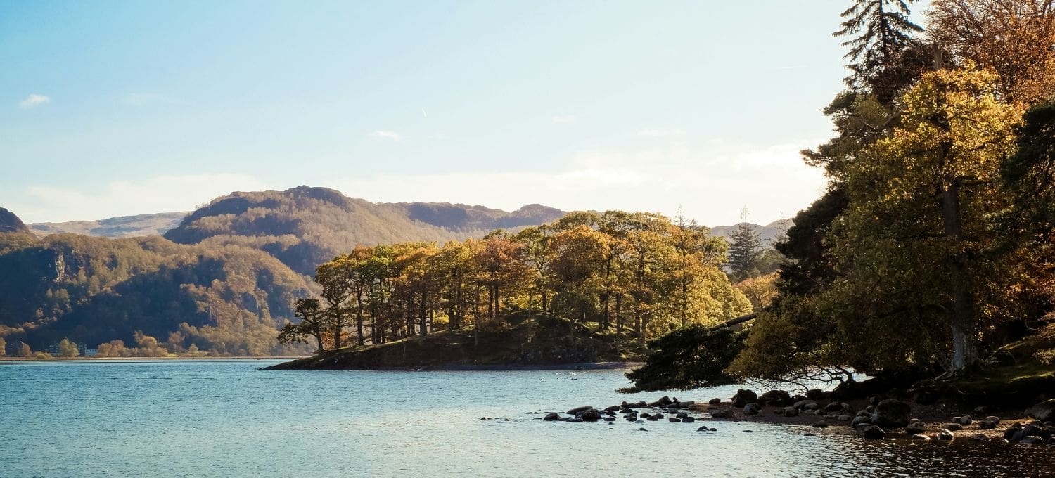 A serene lakeside landscape with a cluster of trees on a hill, surrounded by gentle mountains under a clear blue sky. The scene conveys tranquility.
