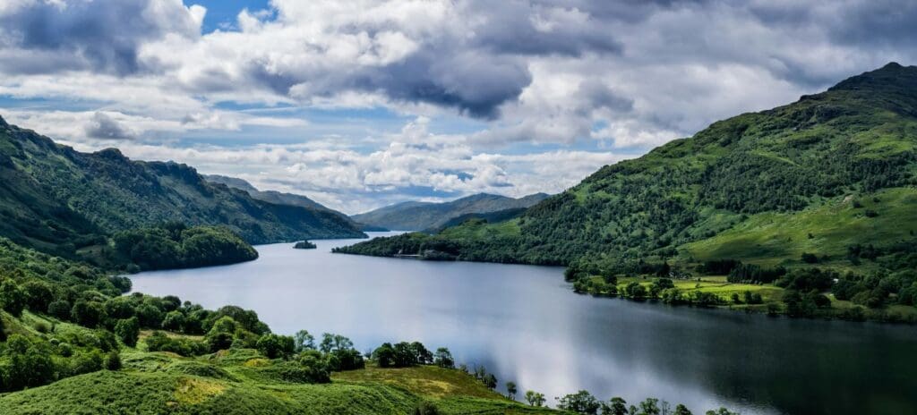 Serene lake with reflective water, surrounded by lush green hills under a partly cloudy sky, creating a peaceful, majestic landscape.