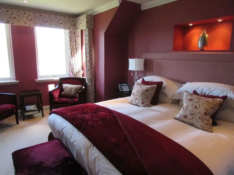 Elegant bedroom with a large bed adorned with white and burgundy pillows and covers. Soft lighting, red walls, accent chair by two windows. Cozy and inviting.