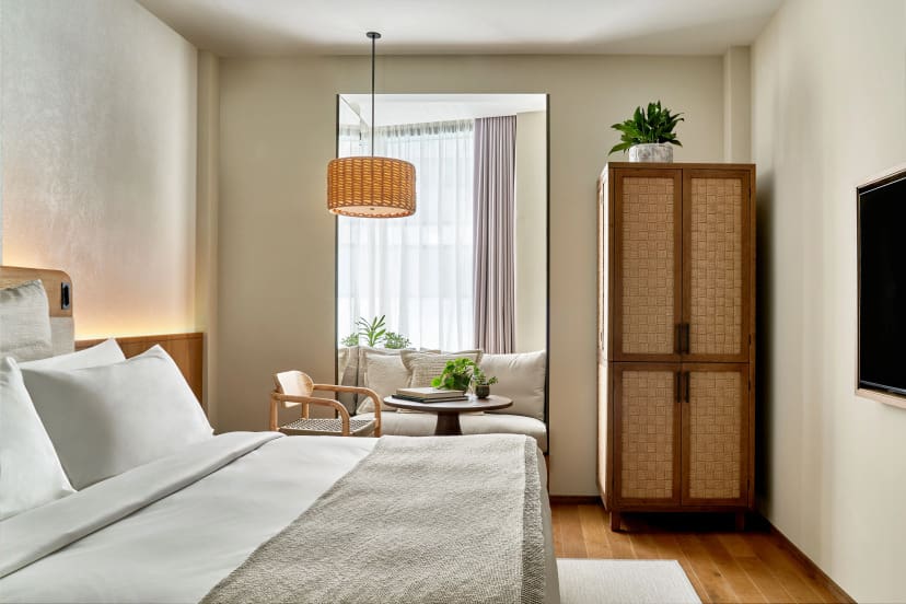 A modern hotel room featuring a king-sized bed, woven pendant light, a cozy seating area with plants, and a stylish storage cabinet.