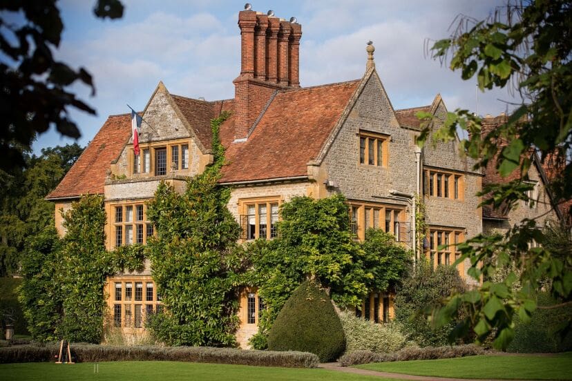 Historic stone manor with ivy-covered walls, tall chimneys, and red-tiled roof. French flag flutters, surrounded by lush greenery and manicured lawn.