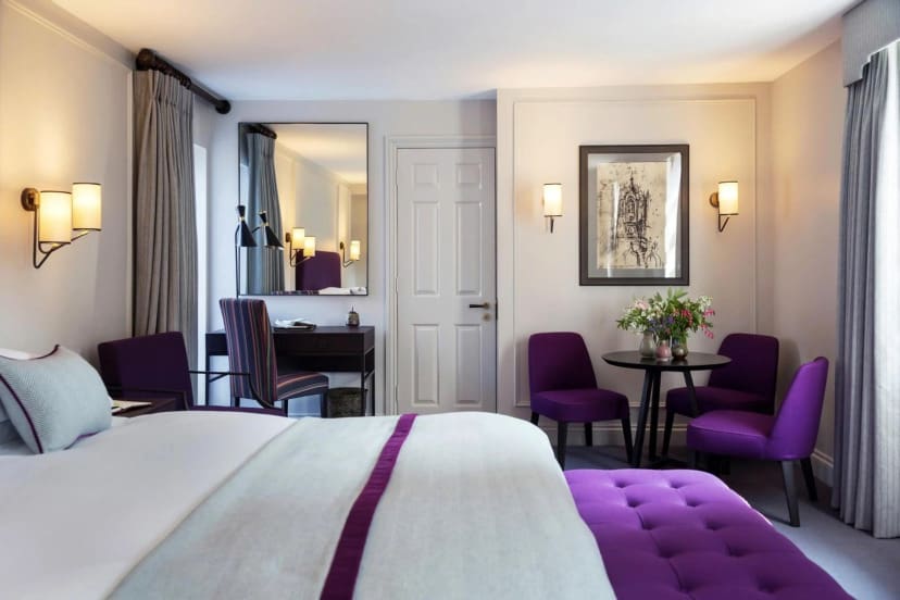 Elegant hotel room with a large bed accented by purple cushions and a striped runner. A small round table with vibrant flowers is surrounded by purple chairs.