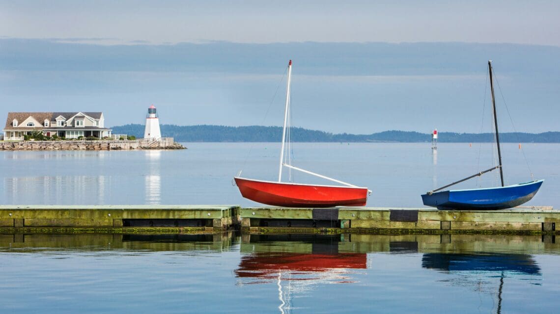 Two sailboats, one red and one blue, are moored at a wooden dock on calm water. A distant lighthouse and house stand on the horizon under a gray sky.