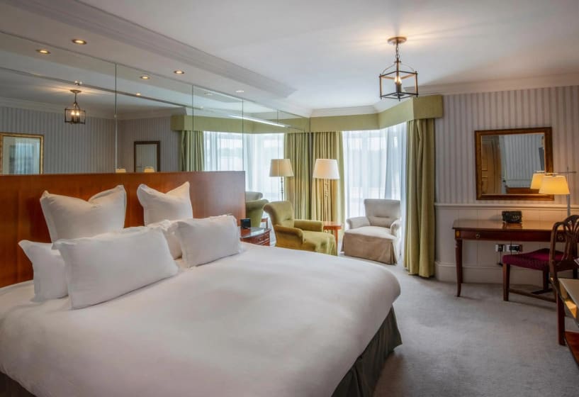 Spacious hotel room with a large bed, white bedding, mirrored headboard, and cozy armchairs by tall windows. Warm lighting and elegant decor.