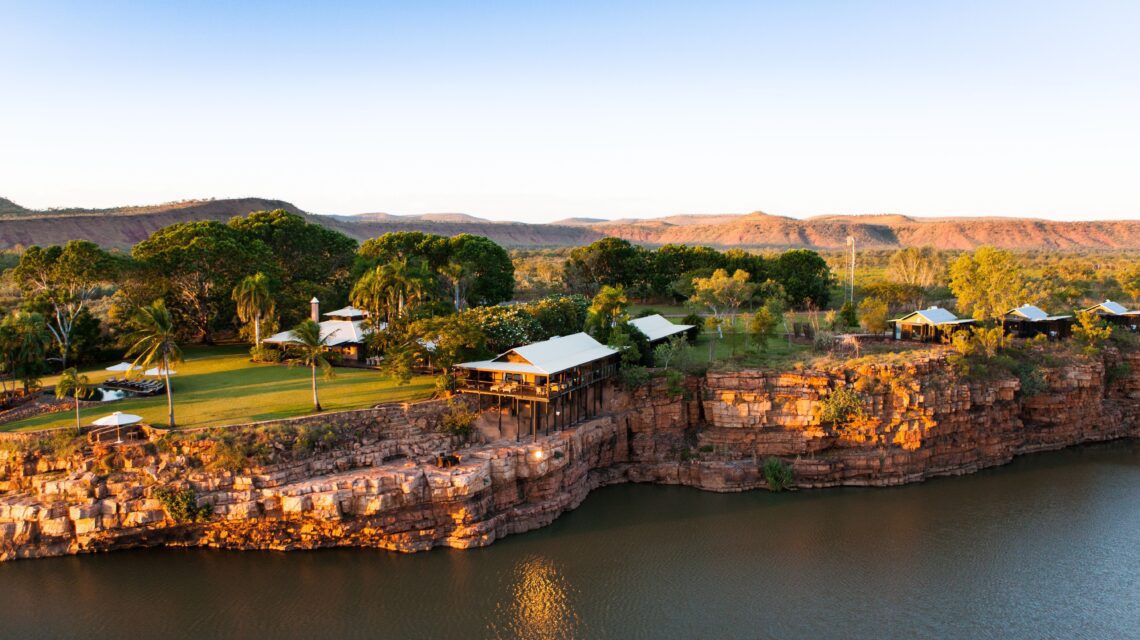 Scenic view of a resort perched on red cliffs beside a calm river. Lush greenery, palm trees, and cabins create a serene, tropical atmosphere.