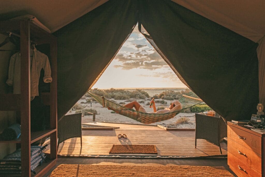 View from inside a tent overlooking a person relaxing in a hammock at sunset. The beach is visible with soft clouds, creating a serene and peaceful atmosphere.