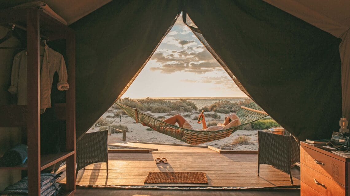 View from inside a tent overlooking a person relaxing in a hammock at sunset. The beach is visible with soft clouds, creating a serene and peaceful atmosphere.