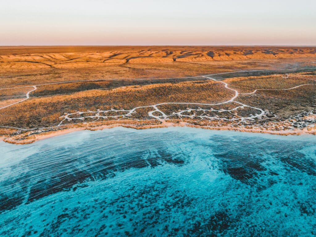 Aerial view of a coastline at sunset, with blue ocean waves meeting the golden-brown sandy land. White winding paths create a pattern on the terrain.