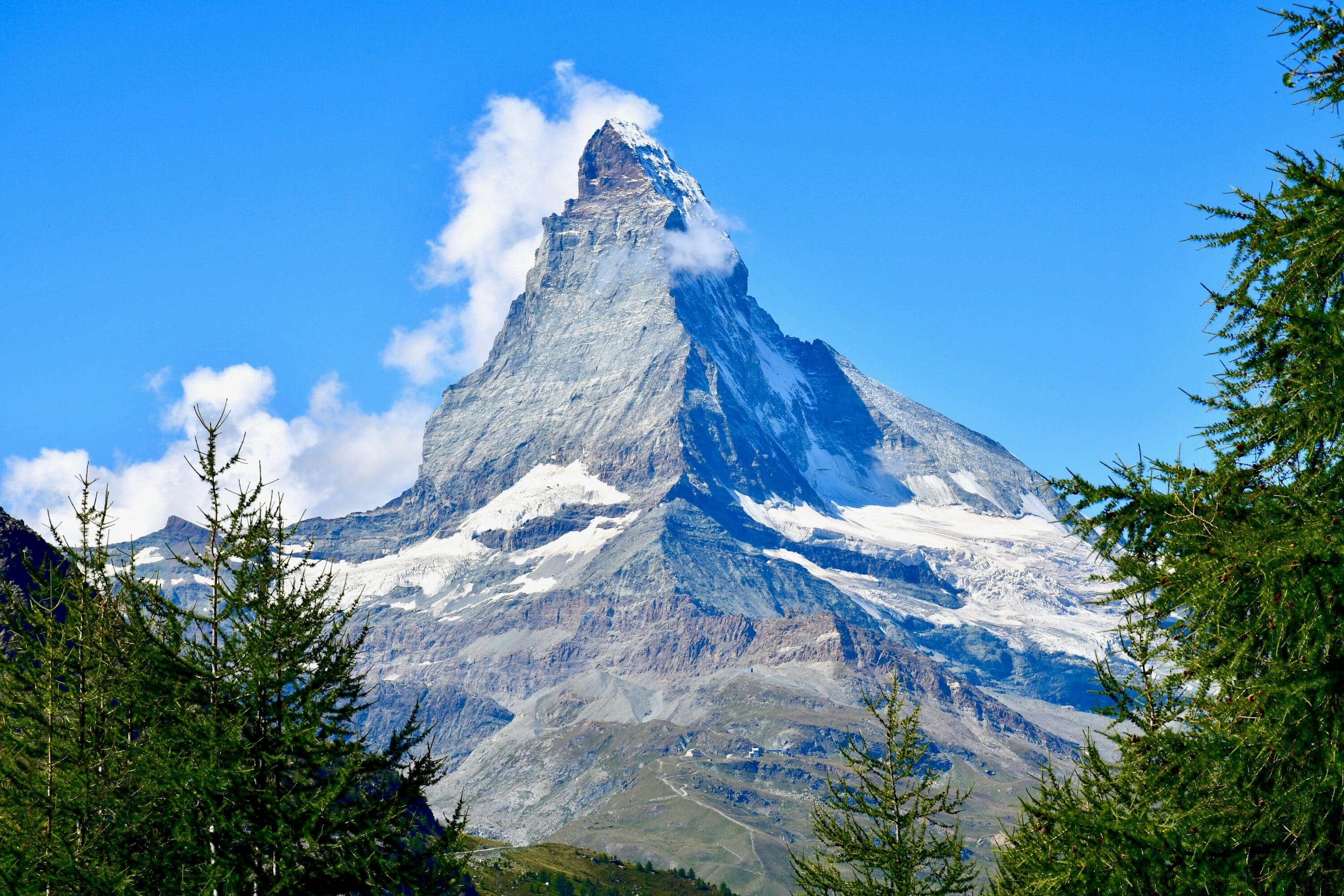 Snow-capped Matterhorn mountain against a clear blue sky, partially veiled by wispy clouds. Foreground features green pine trees. Majestic and serene.