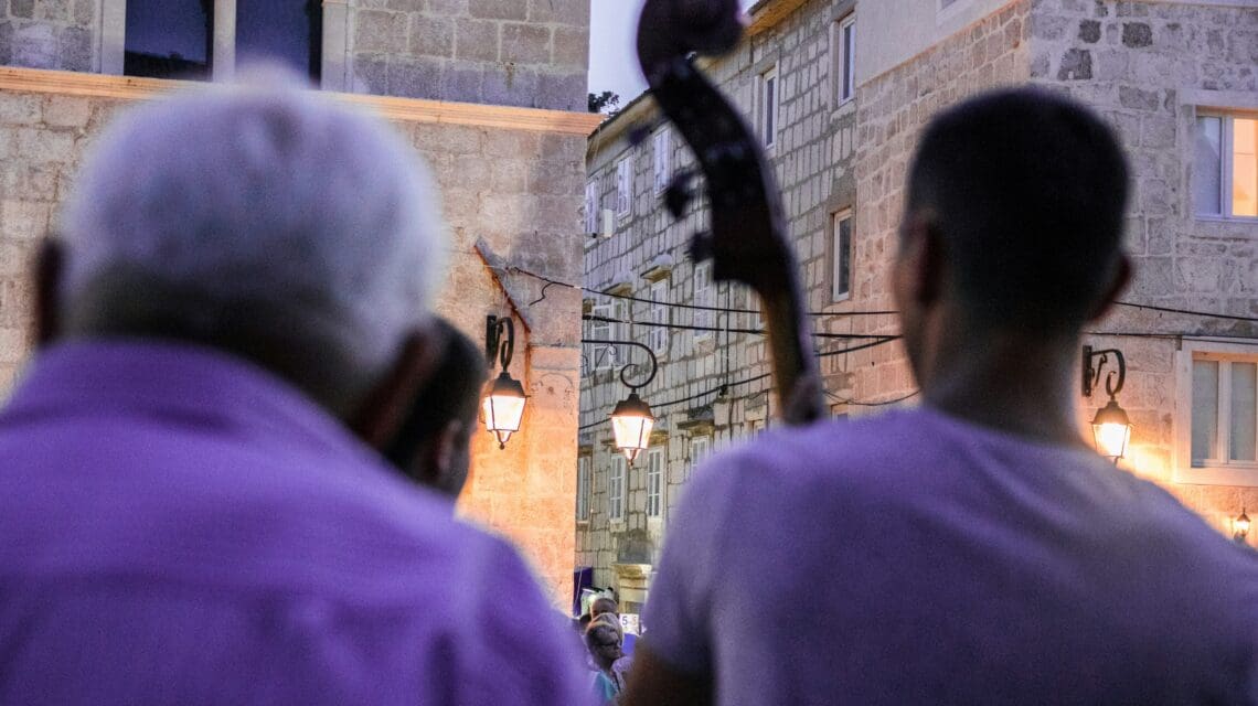 Image of the backs of two people in a white shirts, one holding a standup bass instrument, with string lighs in the background