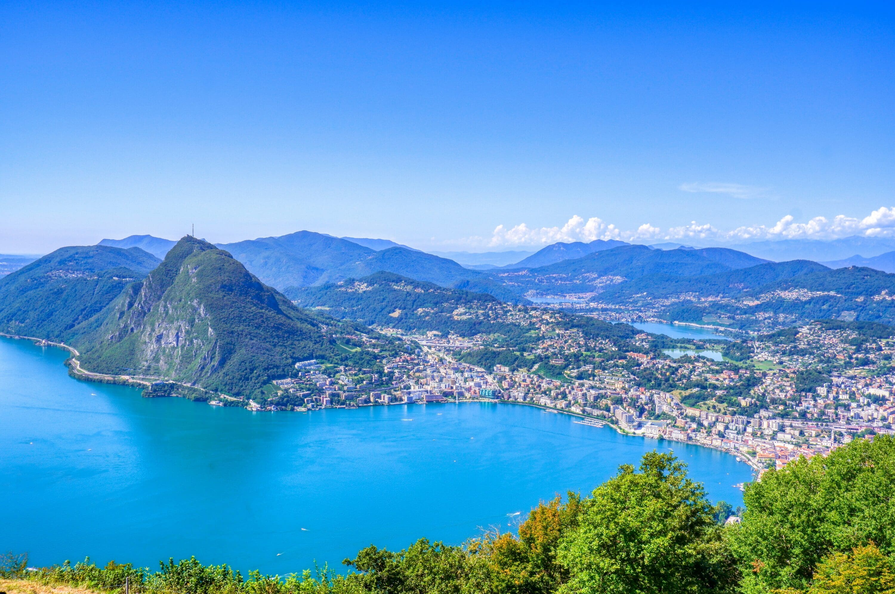 small town next to a lake with a mountain range in the background, few white clouds in the blue sky