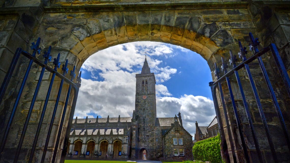 Iron gates opening an archway to a manicured university lawn with a stone tower in the background