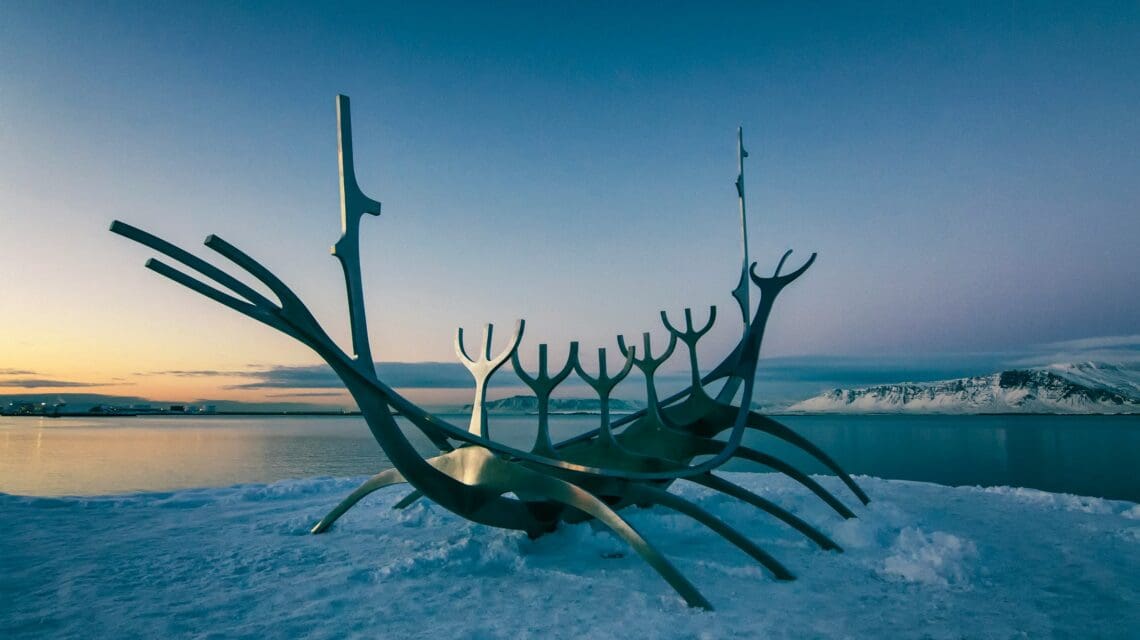 A metallic sculpture resembling a Viking ship sits on snowy ground near calm water at dusk, with a mountainous skyline and a serene, wintry atmosphere.