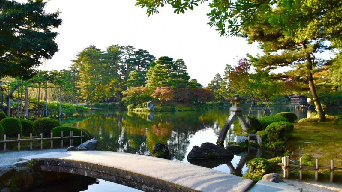 A serene Japanese garden with a stone bridge crossing a reflective pond, surrounded by lush greenery and trees, evoking a peaceful and tranquil atmosphere.